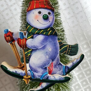 May include: A whimsical Christmas ornament featuring a snowman skiing. The snowman wears a red hat, green and gold scarf, red mittens, and blue boots. The ornament is attached to a small, green Christmas tree.
