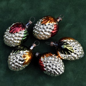 May include: Five vintage glass Christmas ornaments shaped like pine cones. The ornaments have a silver base with colorful accents of red, green, and gold. Each ornament has a small metal loop for hanging.