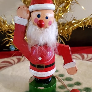 May include: A wooden Santa Claus figurine with a red hat, white beard, and red suit. The figure stands on a green cylindrical base and features articulated arms. A string with a small red wooden bead is attached to the side of the figure.