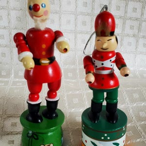 May include: Two wooden Christmas ornaments. One is a Santa Claus figure in a red suit with a black belt and boots. The other is a toy soldier in a red uniform and green pants. Both figures stand on green bases.