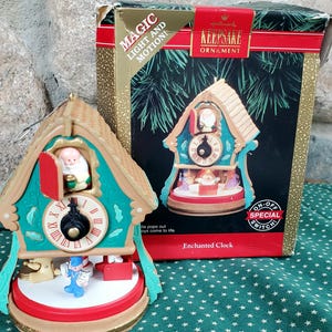 May include: A Hallmark Keepsake Enchanted Clock ornament featuring a cuckoo clock design with a Santa figure in the top door, a central clock face with Roman numerals, and small toy figures on a red base, accompanied by its original product box.
