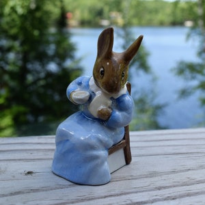 May include: A porcelain figurine of a brown rabbit wearing a blue dress and holding a teacup. The rabbit is sitting on a wooden chair.