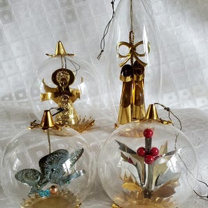 May include: Four clear glass Christmas ornaments with gold accents. Two are spherical, one is oval, and one is a gold angel. The spherical ornaments contain a silver butterfly, red berries, and gold holly. The oval ornament has gold bows.