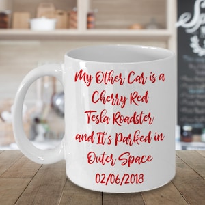 Pode incluir: Caneca de café de cerâmica branca com uma inscrição vermelha que diz "My Other Car is a Cherry Red Tesla Roadster and It's Parked in Outer Space 02/06/2018".
