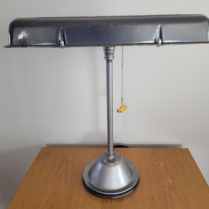 May include: A silver desk lamp with a gray metal shade. The shade is shaped like a car engine valve cover. The lamp has a pull chain switch and a silver base.