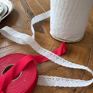 May include: A spool of white eyelet lace and a roll of red ribbon sit on a wooden surface. The white lace has a scalloped edge and small circular cutouts. The red ribbon is tightly wound on a cardboard spool, with a portion unraveled.