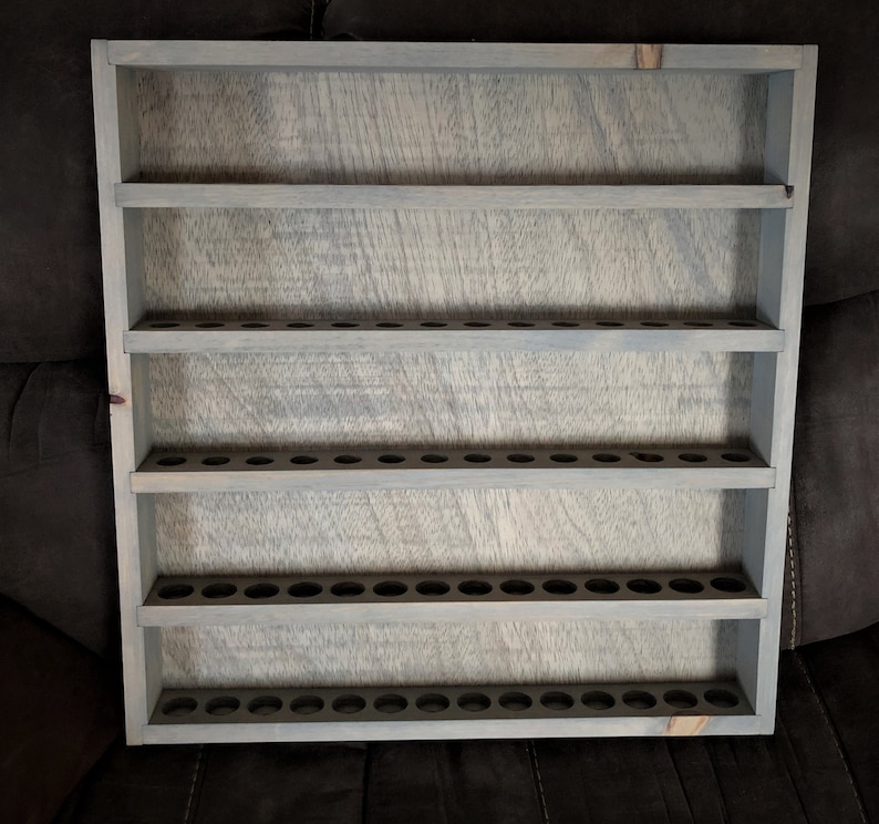 May include: A gray wooden shelf with five shelves and multiple small holes on each shelf. The shelf is designed to hold small bottles or tubes.