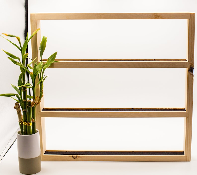 May include: A wooden shelf unit with three shelves. The shelf unit is made of natural wood and has a light brown finish. A potted plant is in front of the shelf unit.