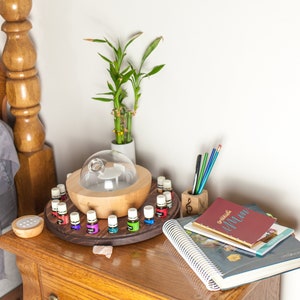 May include: A wooden essential oil diffuser with a glass lid and a rotating tray holding multiple small bottles of essential oils. The diffuser is on a wooden nightstand with a plant, books, and pencils.