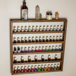 May include: A wooden essential oil rack with five shelves, each holding multiple small bottles with white caps. The rack is brown and has a white background. There is a bottle of rose water spray, a crystal, and a jar on the top shelf.