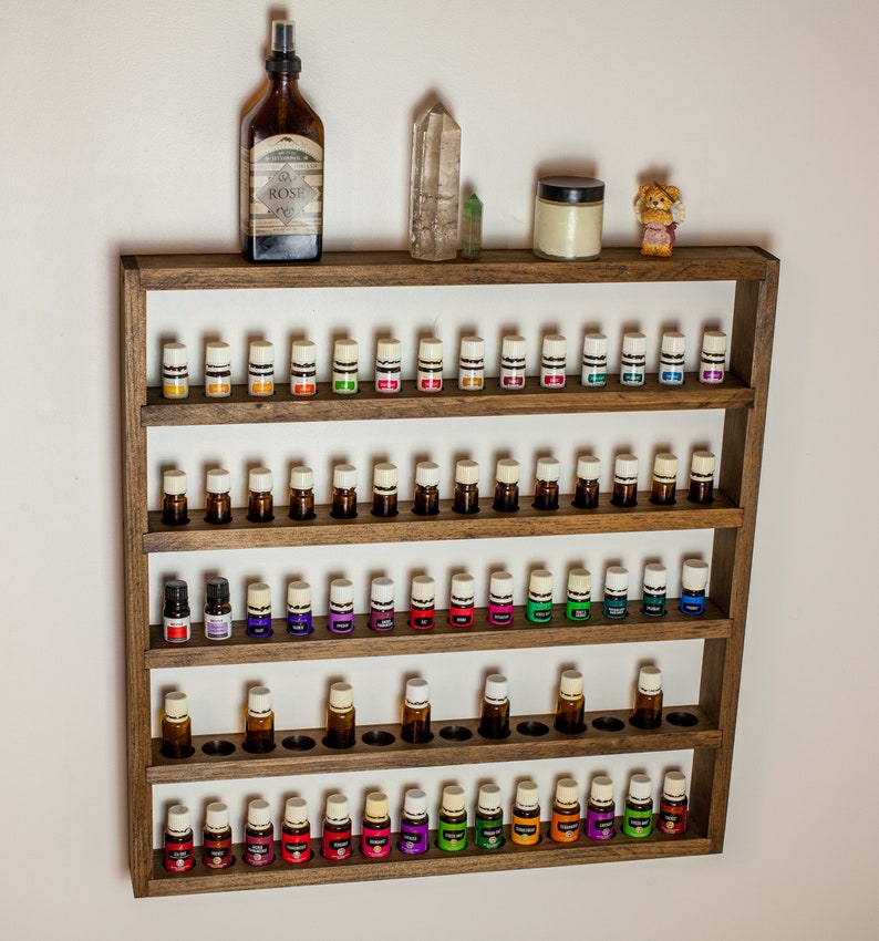 May include: A wooden essential oil rack with five shelves, each holding multiple small bottles with white caps. The rack is brown and has a white background. There is a bottle of rose water spray, a crystal, and a jar on the top shelf.