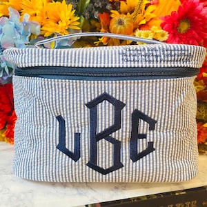 May include: A blue and white striped cosmetic bag with a navy blue zipper and handle. The bag features the embroidered initials "UBE" in navy blue. The background includes a colorful floral arrangement.