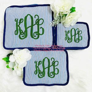 May include: Three blue and white striped travel bags with a dark blue trim. Each bag has a green monogram. White flowers are placed near the bags. The bags are on a white, fluffy surface.