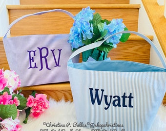 May include: Two seersucker fabric baskets with handles. One basket features purple embroidered initials ERV, and the other features the name Wyatt in dark blue embroidery. The baskets are light blue and white striped with a solid light blue interior.