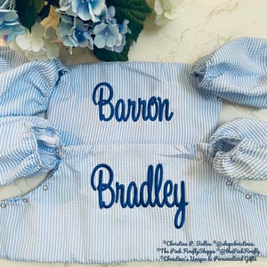 May include: Two light blue and white striped baby outfits with the names "Barron" and "Bradley" embroidered in blue script. The outfits have puffed sleeves and snap closures. Floral arrangement in the background.