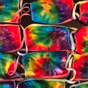 May include: A stack of six colorful tie-dye face masks with adjustable straps. The masks are made of a soft, breathable fabric and feature a variety of vibrant colors, including red, orange, yellow, green, blue, and purple.