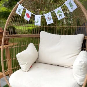 May include: A woven wicker egg chair with a cream-colored cushion and pillows. A banner of illustrated plant cards hangs above the chair. The chair is set outdoors on a patio with artificial grass.