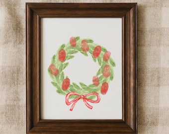 May include: A dark brown wooden frame containing a white print of a green wreath with red fingerprint-style berries and a red painted bow at the bottom. The artwork features a circular arrangement of green foliage with red accents.