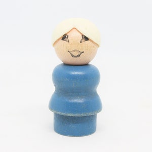 May include: A blue wooden toy figure with a white hairpiece and a painted face. The figure has a smiling expression with black eyes and a pink mouth.