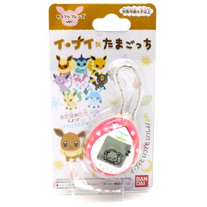 Op de afbeelding: Een roze en witte Tamagotchi-sleutelhanger met een digitaal scherm waarop een slapende Eevee te zien is. De sleutelhanger heeft een zilveren ketting en een kleurrijke illustratie van Eevee en zijn evoluties op de verpakking. De verpakking bevat ook de tekst "イ・ナイ&times;たまごっち", wat zich vertaalt naar "Eevee x Tamagotchi".