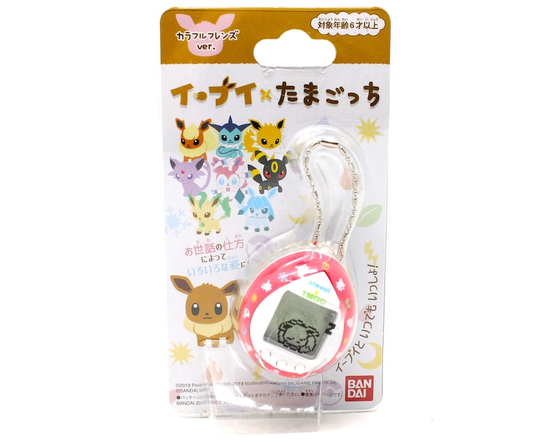 Op de afbeelding: Een roze en witte Tamagotchi-sleutelhanger met een digitaal scherm waarop een slapende Eevee te zien is. De sleutelhanger heeft een zilveren ketting en een kleurrijke illustratie van Eevee en zijn evoluties op de verpakking. De verpakking bevat ook de tekst "イ・ナイ&times;たまごっち", wat zich vertaalt naar "Eevee x Tamagotchi".