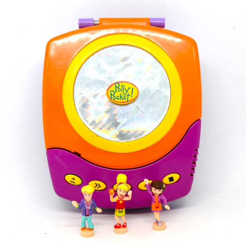 Puede incluir: Un juguete Polly Pocket morado y naranja con forma de reproductor de CD. El juguete tiene una ventana de pl&aacute;stico transparente con un dise&ntilde;o hologr&aacute;fico y las palabras "Polly Pocket!" impresas en &eacute;l. Tres peque&ntilde;as figuras de pl&aacute;stico est&aacute;n de pie frente al juguete.