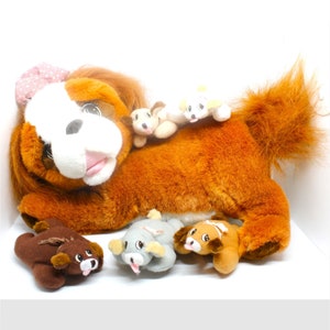 以下が含まれることがあります： ピンクのリボンをつけた茶色のぬいぐるみ犬が、4匹の小さなぬいぐるみの子犬と一緒に横たわっています。子犬は茶色、白、灰色です。