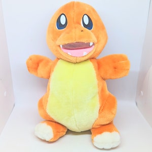 May include: Orange and yellow plush toy of a Pokemon character, Charmander. The toy has a wide smile with white teeth and black eyes.
