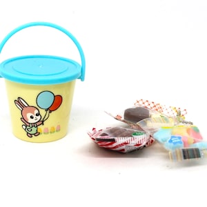 May include: A yellow plastic bucket with a blue lid and handle. The bucket has a cartoon bunny holding balloons printed on the side. The bucket is filled with colorful candies wrapped in clear plastic.