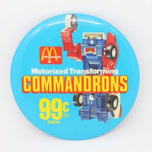May include: A blue and yellow button with a red, blue, and white robot on it. The robot is transforming from a car to a robot. The button says "Motorized Transforming COMMANDRONS 99c EACH".