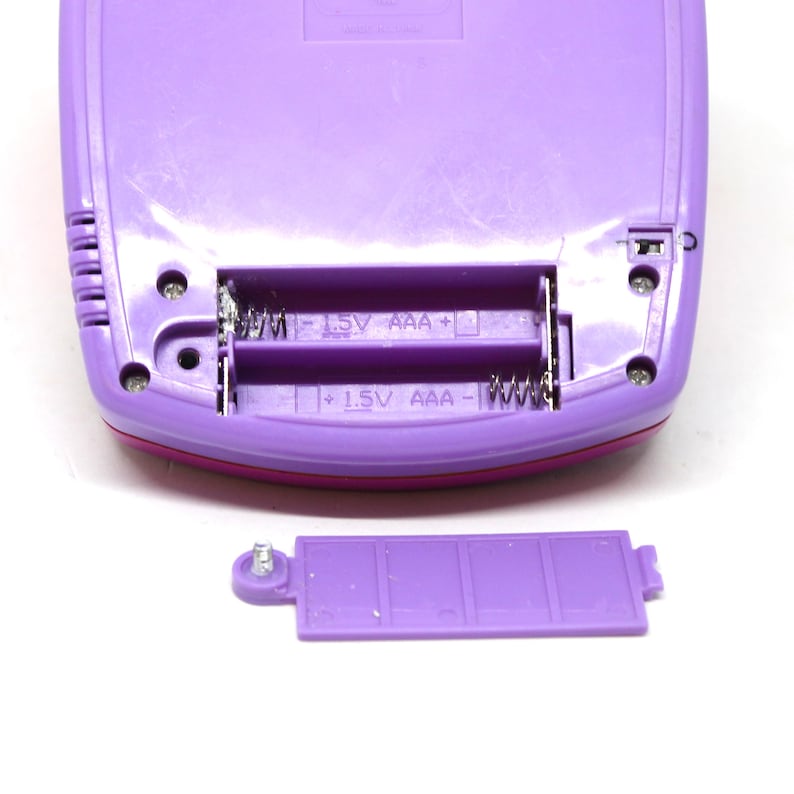 Puede incluir: Compartimento de bater&iacute;a de pl&aacute;stico morado con dos bater&iacute;as AAA y una tapa de bater&iacute;a de pl&aacute;stico morado. El compartimento tiene el texto "1.5V AAA+ 1.5V AAA-" impreso en &eacute;l.