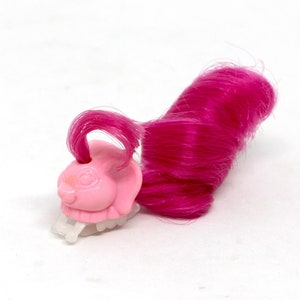 May include: A pink plastic hair clip shaped like a bunny with a tuft of bright pink hair attached. The clip has a clear plastic base with teeth. The bunny has a smiling face and is facing left.