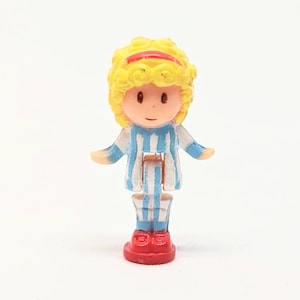 May include: A small plastic doll with blonde hair, a blue and white striped dress, and red shoes. The doll is standing on a red base.