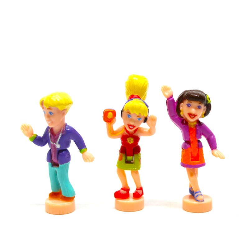 Puede incluir: Tres figuras de pl&aacute;stico de personas, un ni&ntilde;o con cabello rubio que lleva una chaqueta morada y pantalones azules, una ni&ntilde;a con cabello rubio que lleva una blusa roja y una falda verde, y una ni&ntilde;a con cabello casta&ntilde;o que lleva una chaqueta morada y una falda naranja.