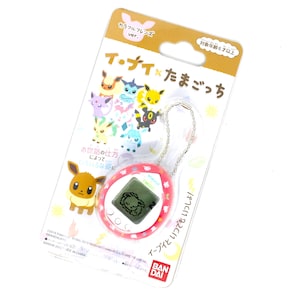 Op de afbeelding: Een roze en witte Tamagotchi sleutelhanger met een digitaal scherm waarop een slapende Eevee te zien is. De sleutelhanger heeft een zilveren ketting en een kleurrijke verpakking met de tekst "イ・ナイ&times;たまごっち" (Eevee x Tamagotchi) en "対象年齢6才以上" (Aanbevolen leeftijd 6 jaar en ouder).