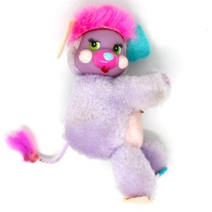 Peut inclure: Un jouet en peluche violet avec de la fourrure rose sur la tête et une oreille bleue. Le jouet a un nez rose et des yeux verts.