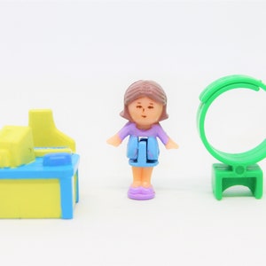 May include: A plastic doll with brown hair, a purple shirt, blue skirt, and purple shoes. The doll is standing next to a yellow and blue desk with a computer on it. There is also a green plastic ring on a stand.