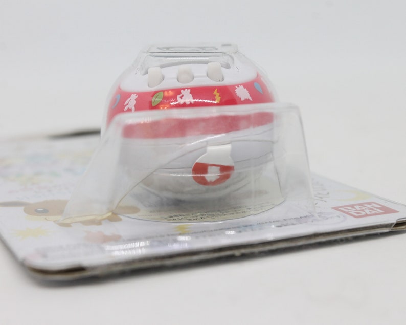 Op de afbeelding: Een rode en witte plastic speelgoedbal met een personage uit de Pokemon-franchise. De bal zit in een doorzichtige plastic verpakking met een witte achtergrond en het woord "BANDAI" erop gedrukt.