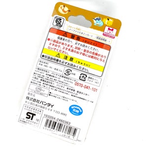 Op de afbeelding: Een gele en bruine kartonnen doos met een afbeelding van een cartoon Eevee en een Tamagotchi op de voorkant. De achterkant van de doos heeft een waarschuwingslabel in het Japans, een telefoonnummer voor klantenservice, een barcode en de tekst "ShoPro 20" erop gedrukt.