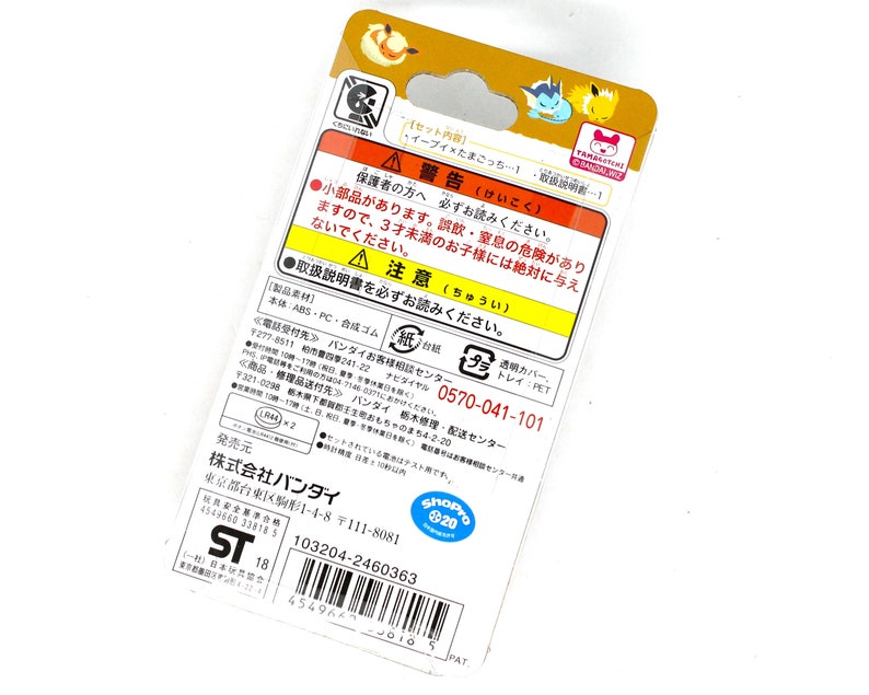 Op de afbeelding: Een gele en bruine kartonnen doos met een afbeelding van een cartoon Eevee en een Tamagotchi op de voorkant. De achterkant van de doos heeft een waarschuwingslabel in het Japans, een telefoonnummer voor klantenservice, een barcode en de tekst "ShoPro 20" erop gedrukt.