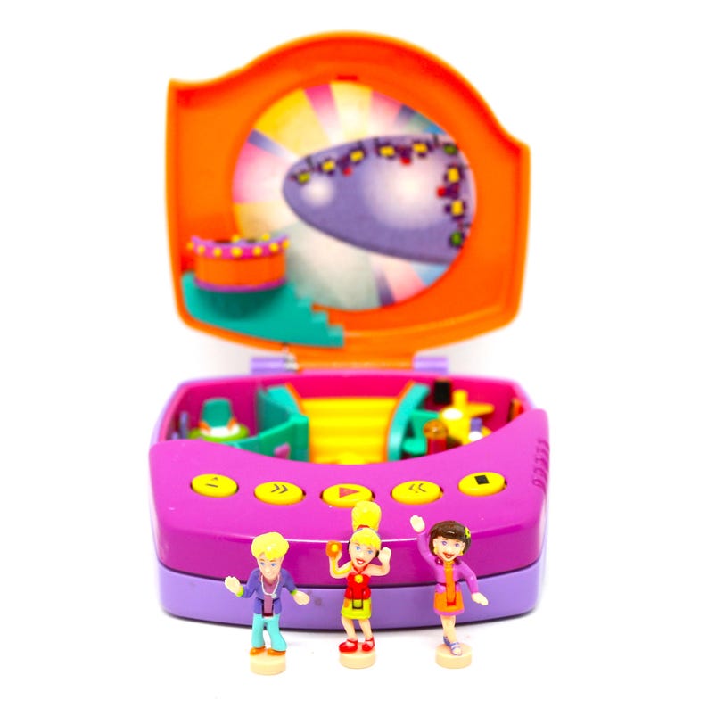 Puede incluir: Un juego de juguetes de pl&aacute;stico morado y naranja con un escenario y tres figuras peque&ntilde;as. Las figuras llevan atuendos coloridos y tienen el pelo amarillo. El juego tiene una base morada con un escenario rosa y un bot&oacute;n amarillo.