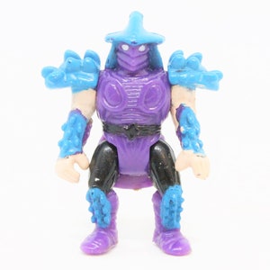 May include: Purple and blue plastic action figure of Shredder from the Teenage Mutant Ninja Turtles franchise. The figure has a spiky helmet and armor.