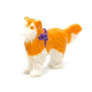 May include: Orange and white plastic toy dog with a purple bow. The dog is standing on a white background.