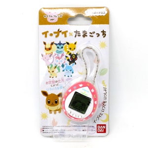 Op de afbeelding: Een roze en witte Tamagotchi-sleutelhanger met een digitaal scherm en een ketting. De sleutelhanger heeft een cartoon-Eevee aan de voorkant en een verscheidenheid aan andere Pok&eacute;mon-personages aan de achterkant. De verpakking bevat de tekst "Eevee x Tamagotchi" in het Japans.