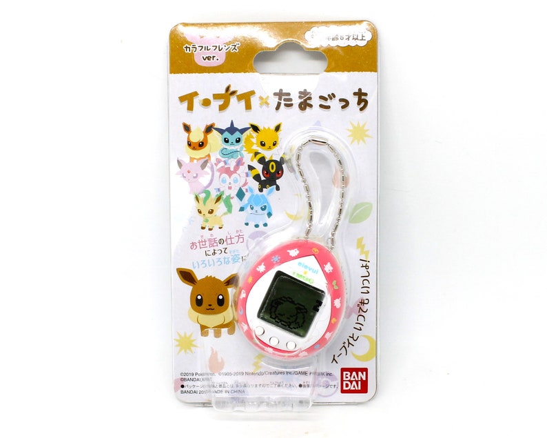 Op de afbeelding: Een roze en witte Tamagotchi-sleutelhanger met een digitaal scherm en een ketting. De sleutelhanger heeft een cartoon-Eevee aan de voorkant en een verscheidenheid aan andere Pok&eacute;mon-personages aan de achterkant. De verpakking bevat de tekst "Eevee x Tamagotchi" in het Japans.