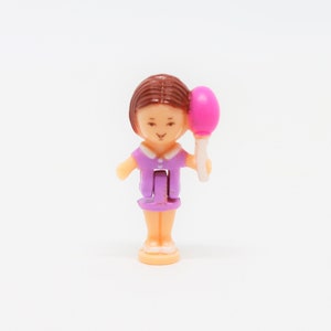 May include: A small plastic doll with brown hair wearing a purple dress and holding a pink balloon.