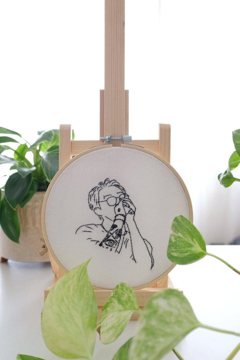 May include: A black and white line drawing of a person wearing glasses and a shirt with a tattoo on their arm. The drawing is embroidered on a white fabric stretched over a wooden embroidery hoop.
