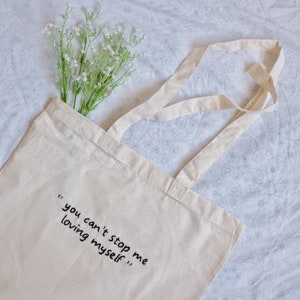 Könnte beinhalten: Eine weiße Stofftasche mit dem Text "you can't stop me loving myself" in schwarzer Tinte.