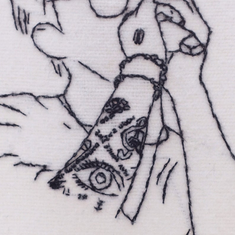 May include: Black and white embroidered image of a person's arm with a detailed eye tattoo. The arm is bent at the elbow and the hand is holding a spray can.