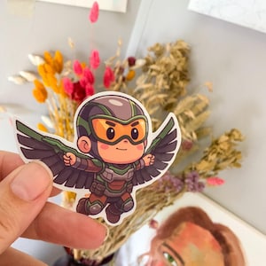 May include: A sticker featuring a cartoon character in a flying pose, with a helmet and wings. The character is wearing a green and brown outfit with orange goggles. The sticker is held in front of a blurred background of dried flowers.
